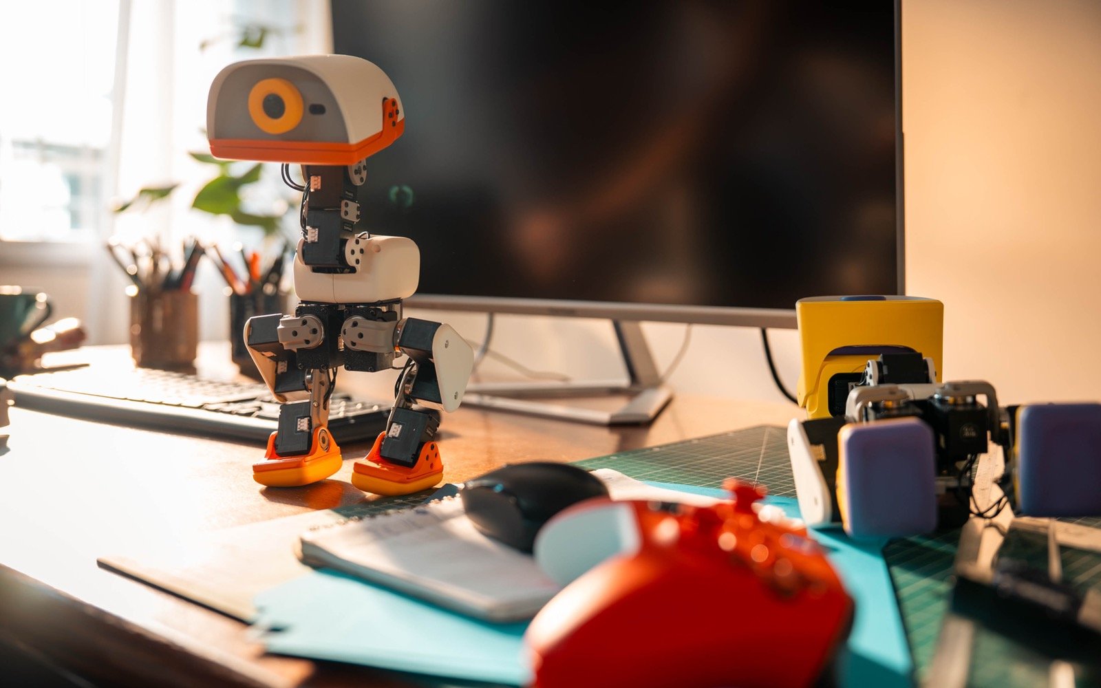 Cream Microduck standing on a wooden desk in morning light, with a monitor, gamepad, and a yellow wheeled accessory nearby.